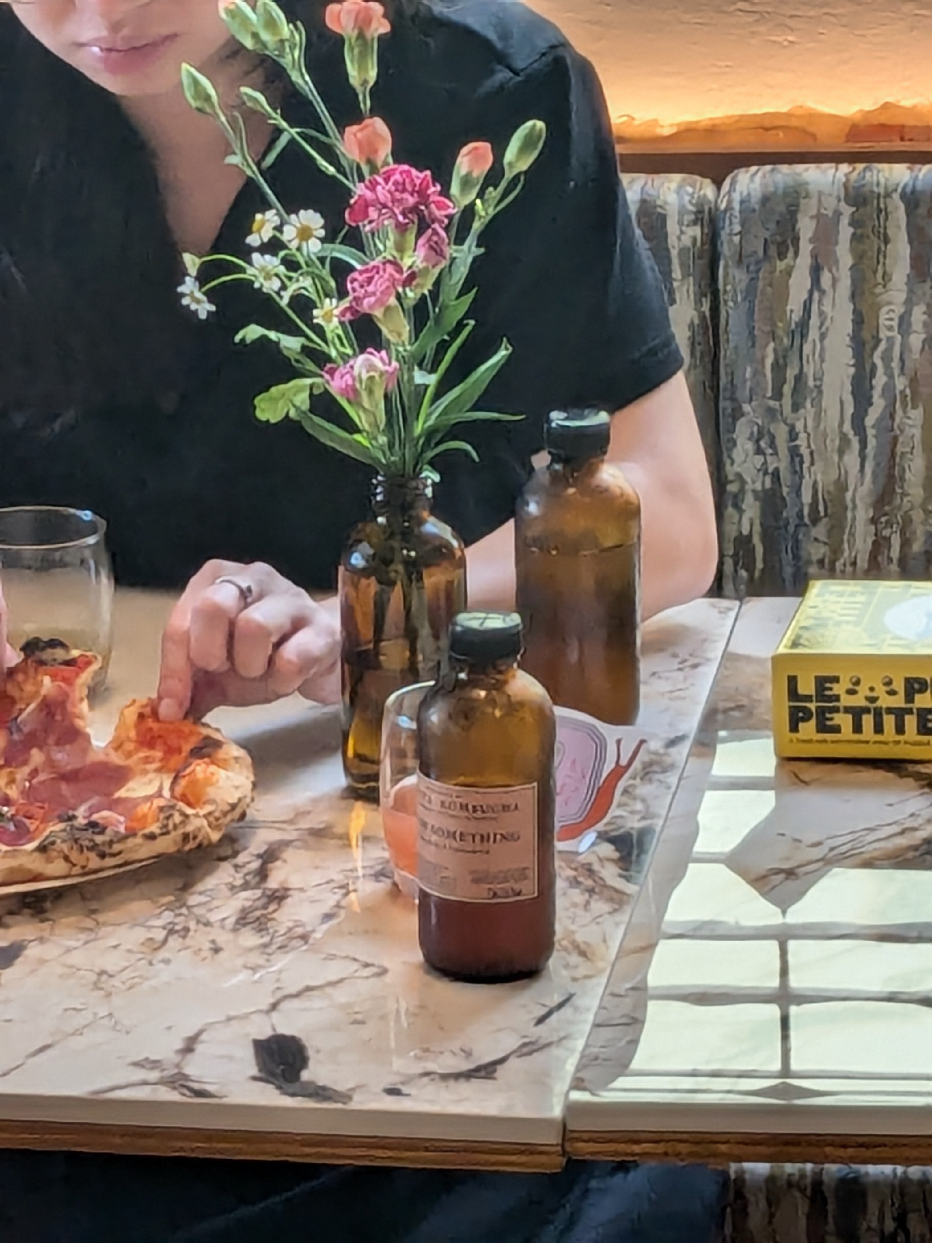 A cozy café scene featuring a marble table with amber glass bottles, one labeled kombucha, a vase with fresh flowers, a partially eaten pizza, and a person in a black shirt reaching for a slice. A yellow box of puzzles titled 'Le Petite' rests on the table.